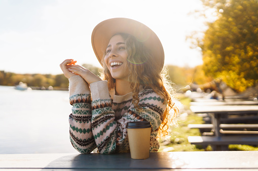 Young tourist woman in a hat drinking coffee and enjoying the sunny weather on a bench by the lake. Concept relax, travel, nature. Active lifestyle