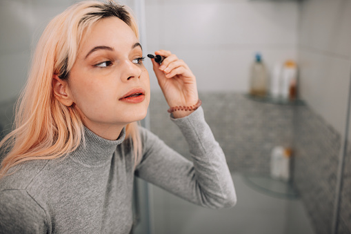 Focused young Caucasian woman applying mascara in bathroom while looking herself in the mirror