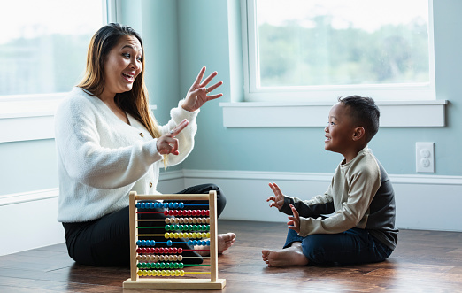 A young, multiracial boy sitting with his mother at home on the floor, learning how to count. They are looking at each other, holding up fingers. An abacus is on the floor next to them. Mother is an Asian woman in her 30s, and her son is mixed race, Asian and African-American, 5 years old.