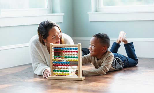 A young, multiracial boy lying with his mother at home on the floor, learning how to count with an abacus. Mother is an Asian woman in her 30s, and her son is mixed race, Asian and African-American, 5 years old. They are smiling and looking at each other.