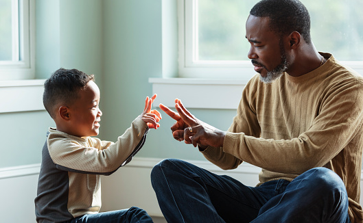 A young mixed race boy sitting with his father at home, on the floor. They are facing each other, holding up fingers, playing a little game. Father is a mature African-American man, in his 40s. His son is African-American and Asian, 5 years old.