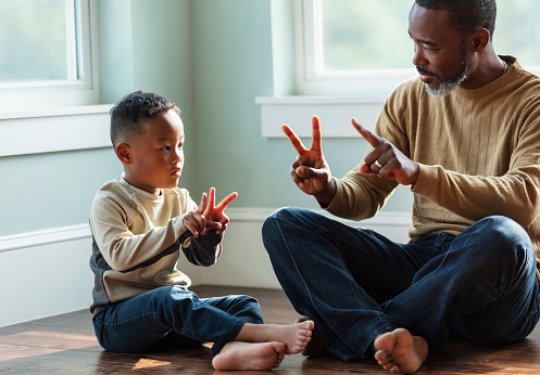 A young mixed race boy sitting with his father at home, on the floor. They are facing each other, holding up fingers, playing a little game. Father is a mature African-American man, in his 40s. His son is African-American and Asian, 5 years old.