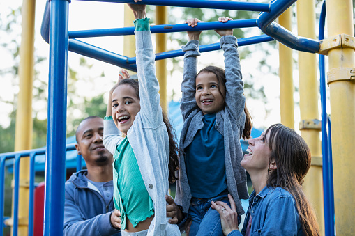 A multiracial family having fun playing at the playground. The two daughter are hanging from monkey bars. The girls, 7 and 9 years old, are mixed race, Caucasian, African-American, and Hispanic. Mother and father are standing below, looking up and helping them. The older girl is looking toward the camera.