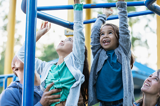 A multiracial family having fun playing at the playground. The two daughter are hanging from monkey bars. The girls, 7 and 9 years old, are mixed race, Caucasian, African-American, and Hispanic. Mother and father are standing below, looking up and helping them.