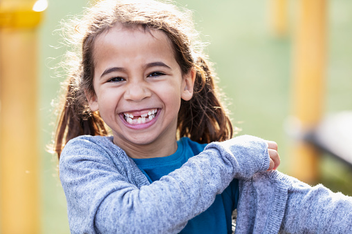 Headshot of a mixed race 7 year old girl with a big toothy smile, grinning at the camera. She is outdoors, at the playground on a sunny day.