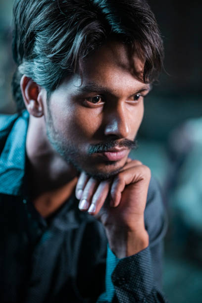 An Asian/Indian young man sits at home, deep in thought, resting his chin on his hand.