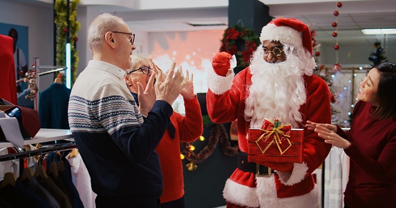 Retail assistant wearing Santa Claus suit in holiday adorn fashion boutique, holding present box acting as raffle tickets container, drawing random senior woman among happy clients