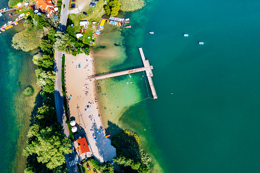 A beach and a pier on a green lake in Kashubia, Poland. The photo was taken from above, giving a bird’s eye view of the beach and the pier. The beach is sandy and has a few umbrellas and people on it. The pier is wooden and extends into the lake. The lake is green and has a few boats on it. The shore has a few houses and trees on it. The image is taken during the day and the colors are bright and vibrant. The photo shows the fun and relaxing atmosphere of the beach and the pier.