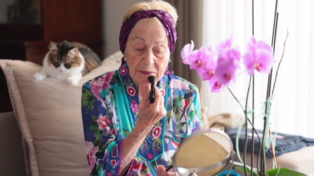 Beautiful senior woman doing makeup in front of mirror and her cat at home.