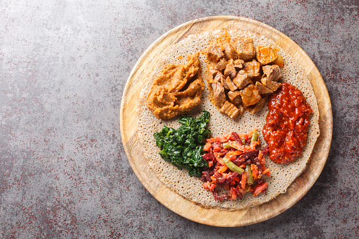 Ethiopian Injera topped with meat, vegetables, greens close up on the wooden board on the table. Horizontal top view from above
