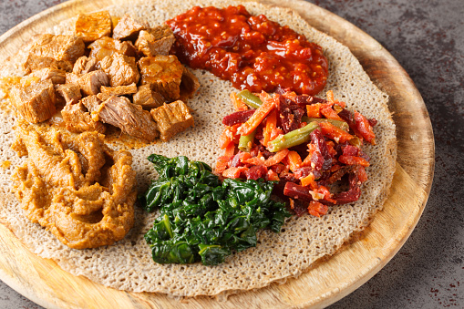 Ethiopian Injera topped with meat, vegetables, greens close up on the wooden board on the table. Horizontal