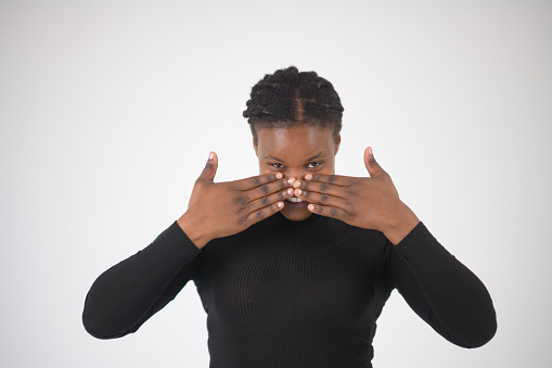 Beautiful Smiling African woman studio portrait peeking at camera above hands held up.  Her hands are facing outwards close to her nose obscuring her smile.