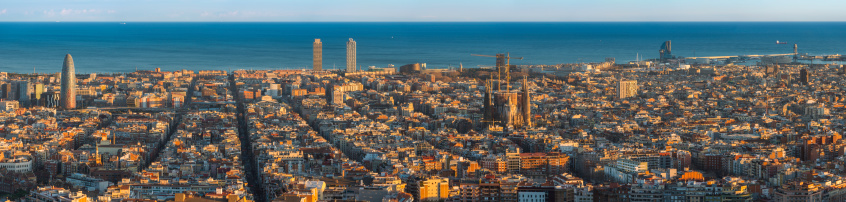Golden sunset light illuminating the landmarks and avenues of Barcelona, from the futuristic curves of the Torre Agbar, along the blue waters of the Mediterranean shore past the Torre Mapfre and Hotel Arts to the iconic spires of Gaudi's Sagrada Familia and the quayside developments of Port Vell at the end of La Rambla and the Barri Gotic, Catalonia, Spain. ProPhoto RGB profile for maximum color fidelity and gamut.