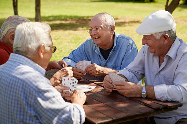 Group of senior men, smiling and playing cards at the park Active retirement, old people and seniors free time, group of four elderly men having fun and playing cards game at park. Waist up park bench photos stock pictures, royalty-free photos & images