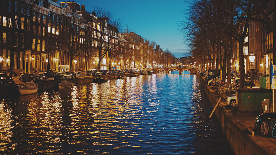 Amsterdam's enchanting nightscape unfolds beneath an arch bridge,spanning a serene canal. The scene is adorned with bare trees,casting delicate silhouettes against a row of glowing street lights. The residential buildings stand as witnesses to the city's timeless allure.