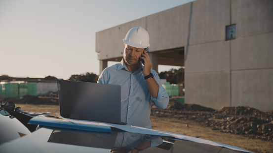 Mature male building contractor balances his task and examining laptop while discussing over smart phone at busy construction site