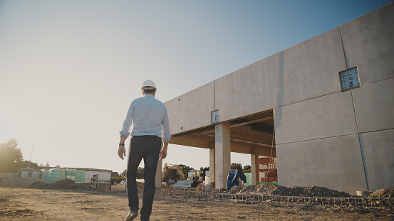 Confident male project manager with protective headwear walking towards progressive under construction building during sunset