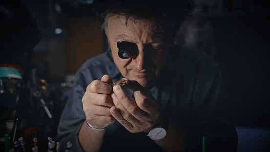 A mature male watch maker wears a magnifying glass while repairing the gear of a wristwatch at a repair shop