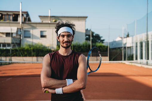 Portrait of a tennis player on the field. He's looking at camera, holding a racket and a tennis ball.