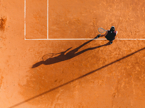 Aerial view of a tennis player during a match. Drone shot directly from above. Orange tennis court field.