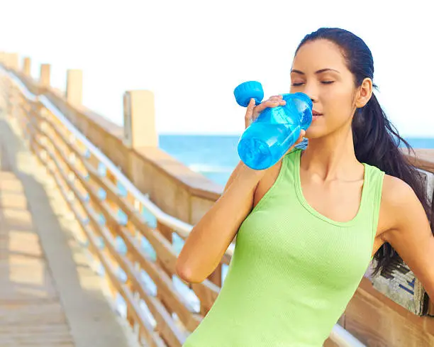 Healthy young woman drinking from waterbottle on boardwalk. Horizontal shot.