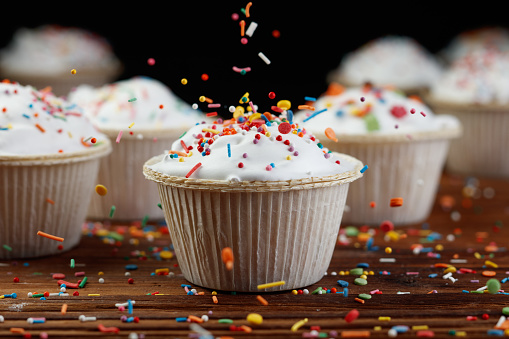 Сupcakes or muffins decorated with confectionery decor on a wooden background. Сonfectionery confetti, balls flying in the air. Shallow depth of field