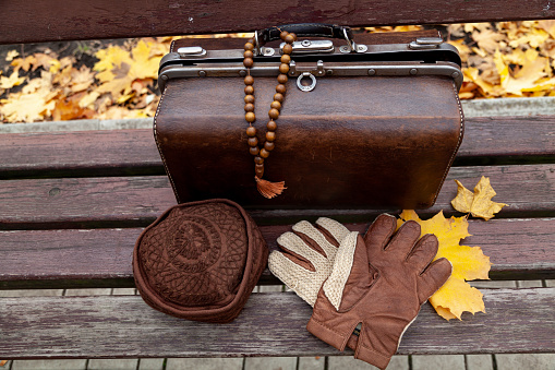 Still life with a valise and a rosary. There is a leather valise on a wooden bench in the park. There is a wooden rosary on it. Nearby lies a hat and gloves