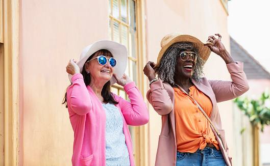 Two multiracial senior women taking a vacation together, sightseeing in town. The African-American woman is in her 70s. Her Caucasian friend is in her 60s. They are smiling, looking up, wearing sunglasses and wide brimmed sun hats.