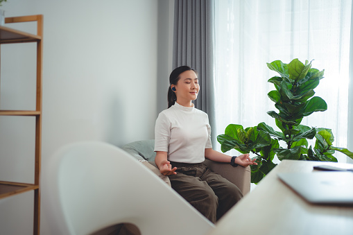 Asian woman calms down taking a break with meditating holding her hands in yoga gesture sitting at home office. Happy woman working from home.