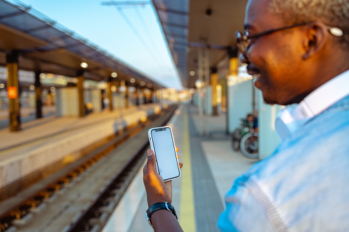 Shot of the African American young man waiting for a train at a railroad station. He is using smart phone with blank screen