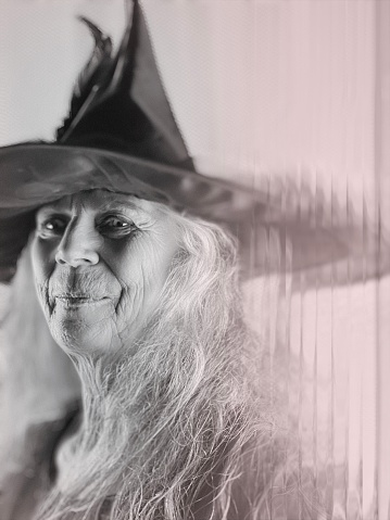 Cute senior woman, with long silver hair, dressed as a witch for Halloween, she is wearing a big witch hat. She is looking away from the camera. Defocused background with a wave pattern and some slight pink lighting.