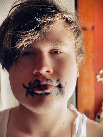 Cute teen boy, posing inside his home, while getting ready for a party on Halloween night. He is wearing black face make up that looks like his mouth is criss crossed with fake stitches.