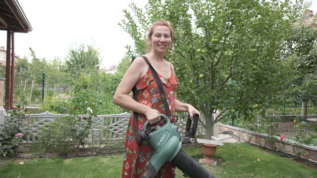Slow motion of Mature woman posing for camera while cleaning leaves using leaf blower in backyard.