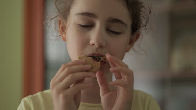 Little Girl Eating a Cookie. Looking at Camera and Smiling. Cute Child Hungry Eating Cookie Enjoying Delicious Treat at Home in Kitchen. Junk Food, Unhealthy Nutrition Sugar.