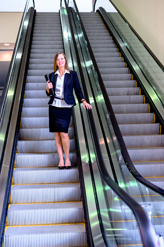 istock Professional woman in her mid-50’s with file folder on an escalator in a large office building or corporate campus 1623587425