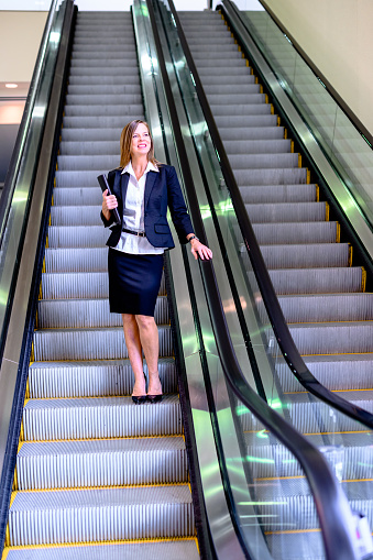 istock Professional woman in her mid-50’s with file folder on an escalator in a large office building or corporate campus 1623587548