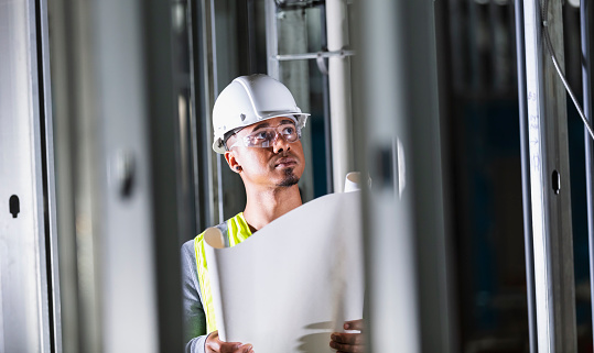 A mid adult multiracial man at a construction site wearing a white hardhat, holding a set of blueprints, and examining the structure being built. He is in his 30s, black and Hispanic. He could be a foreman, engineer, or perhaps a building contractor or safety inspector.