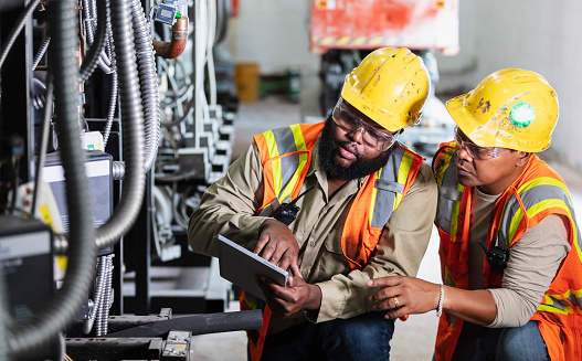 Two multiracial men inspecting the electrical components of an industrial refrigeration system. They are wearing hardhats, safety glasses and safety vests. The mid adult man holding the digital tablet is African-American, in his 30s. His coworker is black, Hispanic and Asian, in his 40s.