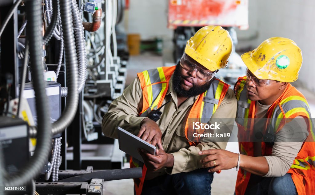 Two workers inspecting industrial refrigeration equipment Two multiracial men inspecting the electrical components of an industrial refrigeration system. They are wearing hardhats, safety glasses and safety vests. The mid adult man holding the digital tablet is African-American, in his 30s. His coworker is black, Hispanic and Asian, in his 40s. Electrician Stock Photo