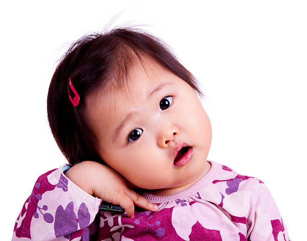 Cute Asian baby, holding a cellular phone to her ear and looking at the camera, isolated on white.  Baby is 15 months old and has her head cocked to the side.