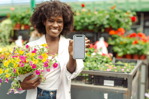 A young African American female florist is holding flowers and showing her phone, while standing in front of her market stall. She's cheerfully looking at the camera.