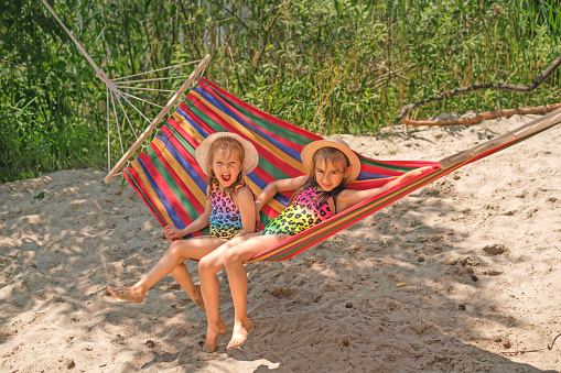 Laughing funny tanned girls in swimsuits and hats relax in a hammock on the beach.  Summer holidays on vacation