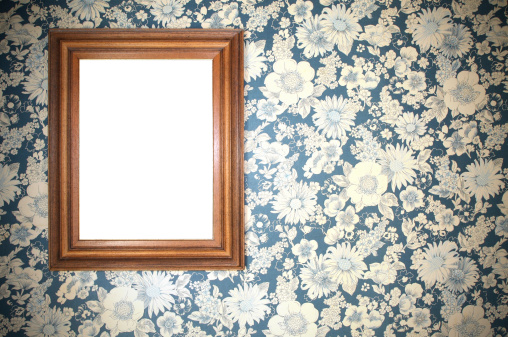 Old wooden picture frame, blank, on a wall, decorated with an old floral wallpaper discolored 1970's, with white flowers turned yellow with time. Wallpaper juncture visible on the right of the image ... Vignetting on the edges of the photo.