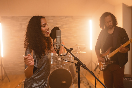 Passionate black female musician enjoying singing in the studio with her band. She is passionately singing into the microphone and waving her hands around looking down. Her hair is dark and curly and her clothes are silver and sparkly. She is accompanied by her bandmates. A curly haired guitarist is playing his electric guitar while the drummer is playing the drums in the back.