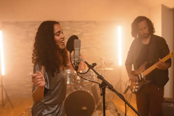 Passionate black female musician enjoying singing in the studio with her band. She is passionately singing into the microphone and waving her hands around looking down. Her hair is dark and curly and her clothes are silver and sparkly. She is accompanied by her bandmates. A curly haired guitarist is playing his electric guitar while the drummer is playing the drums in the back.