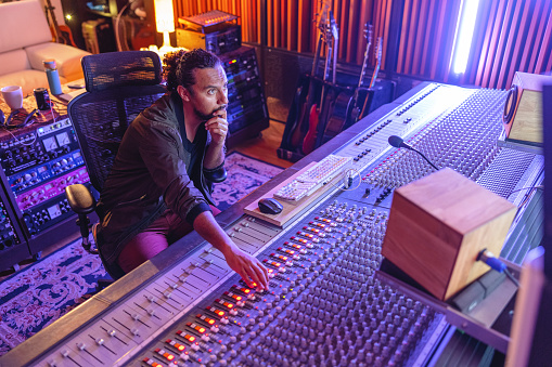A portrait of a black male sound engineer producing a song in a cool recording studio. He is sitting on a chair and making adjustments to the sound on a large music mixer. The dark studio is illuminated by orange and violet led lights. The male sound engineer is attractive and has a beard and long curly hair. There is music equipment surrounding the man.