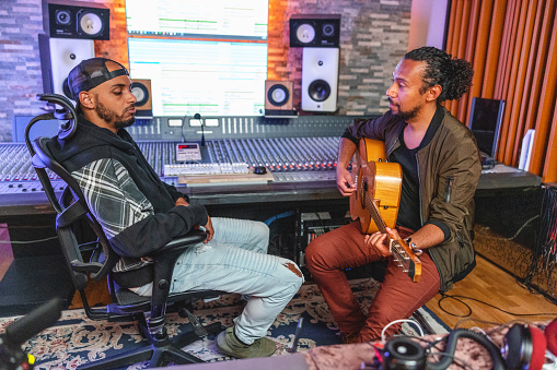 An adult male black long curly haired musician playing the acoustic guitar for a cool bearded male music producer in a recording studio. They are surrounded by tons of music and recording equipment including loudspeakers and a music mixer. They look serious.