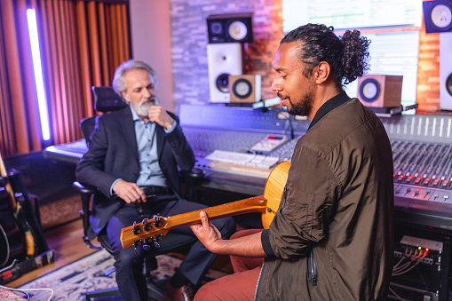 An adult black male guitarist playing an acoustic guitar for a senior caucasian producer with grey hair. The producer is focused on listening to the song and wearing a classy dark suit. They are surrounded by music equipment, loudspeakers and an audio mixer.