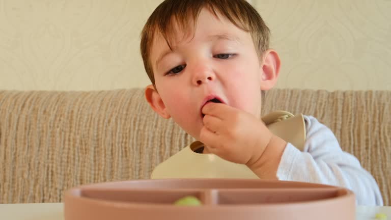 The baby is happily eating fruits and berries, sitting at the table with a bib on. Kid boy aged two years (two-year-old)