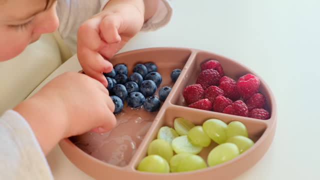 The child is happily munching on some fruits and berries while sitting at home. Kid boy aged two years (two-year-old)